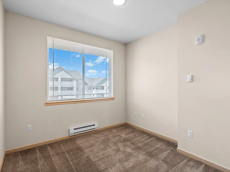 Bright room with plush carpeting and large window offering a picturesque view of neighboring buildings and blue sky. Bright room with plush carpeting and large window offering a picturesque view of neighboring buildings and blue sky.