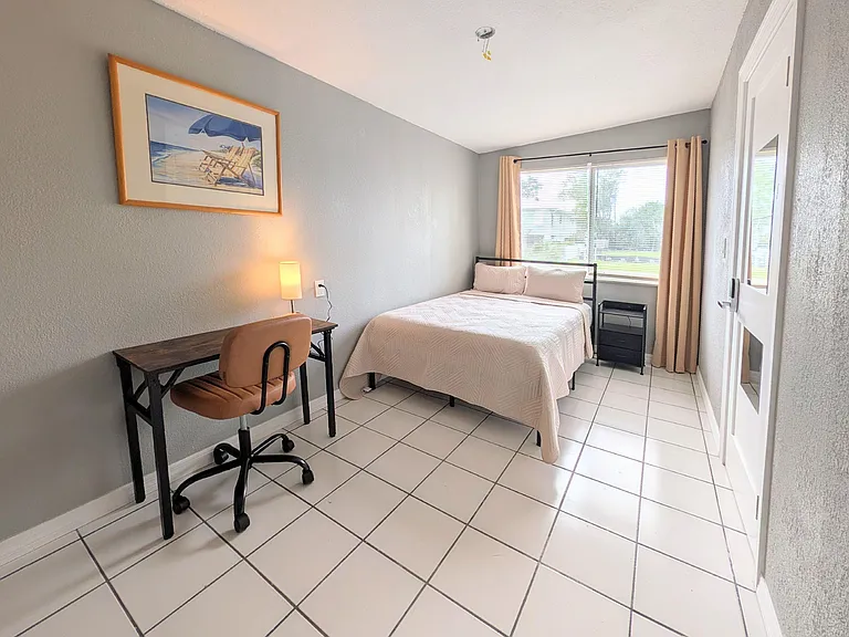 Private furnished bedroom with a full bed, work desk, and nightstand, plus a large window with curtains for natural light.