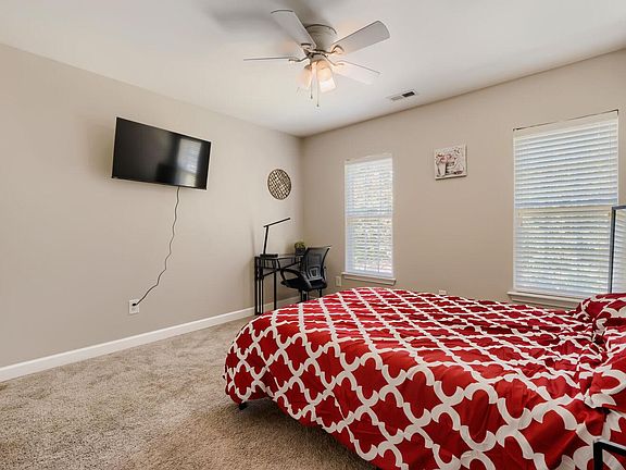 Spacious bedroom with two windows, ceiling fan, wall-mounted TV, and a dedicated desk area on soft carpet.