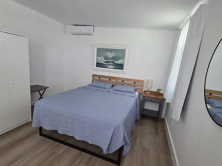 Furnished bedroom with queen bed, nightstand, closet storage, and window with curtains plus wall-mounted A/C.