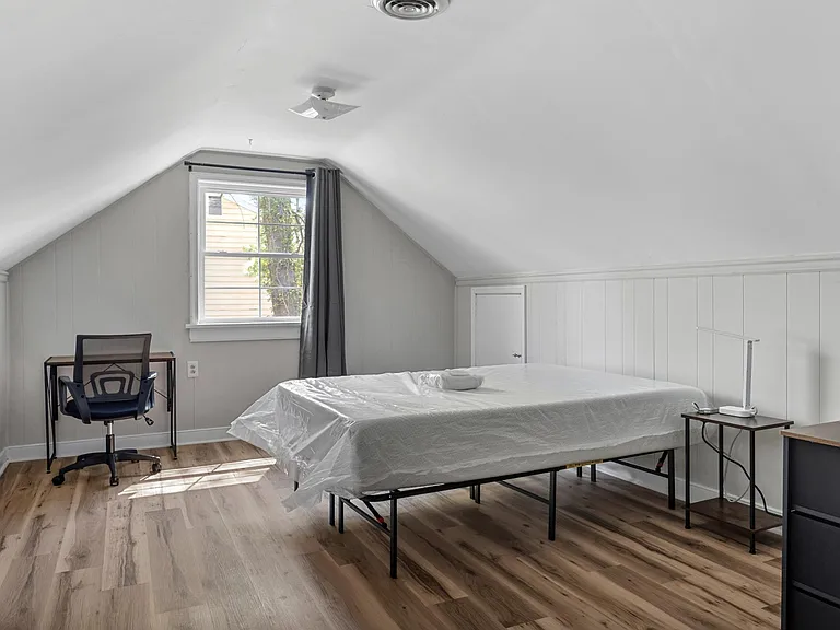 Upper-level bedroom with sloped ceilings, wood-look flooring, a work desk, and a window with curtains for natural light.
