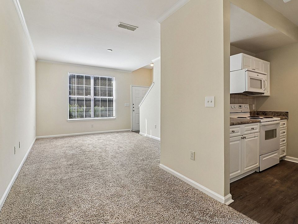 Bright and inviting open-plan living area with plush carpet, large window, and compact modern kitchen featuring white cabinetry.