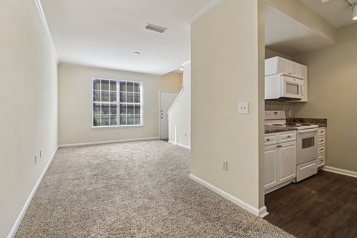 Bright and inviting open-plan living area with plush carpet, large window, and compact modern kitchen featuring white cabinetry.