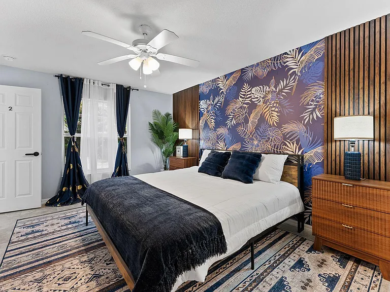 Spacious furnished bedroom featuring a queen bed, matching nightstands, ceiling fan, and window with layered drapery.
