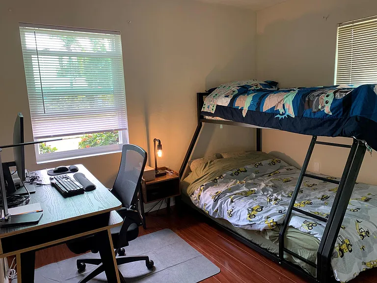 Second bedroom with a bunk bed (full at the bottom and twin on the top) and an office desk with monitor, lamp, keyboard, mouse, phone charger, and a chair.