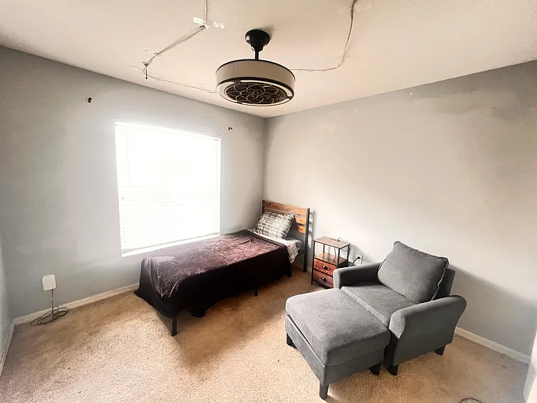 Furnished bedroom with a twin bed, nightstand, and lounge chair, plus a large window with blinds and soft carpet.