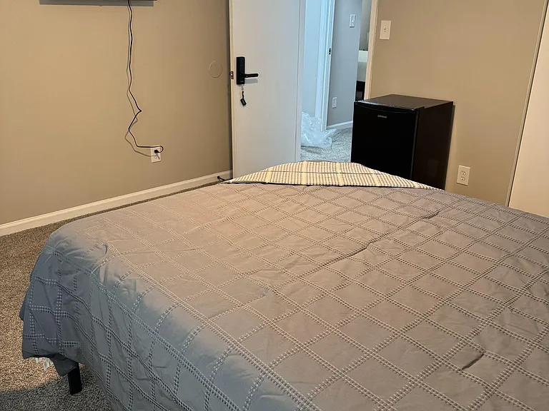 Furnished private bedroom with a comfortable bed, wall-mounted TV, and mini fridge for added convenience.