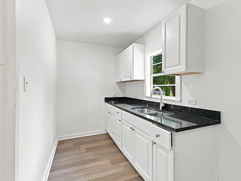 Galley kitchen featuring white cabinetry, dark stone countertops, a stainless steel double sink with a gooseneck faucet, and wood-finish flooring