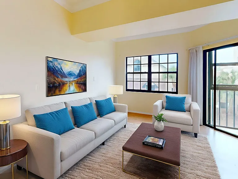 Bright and inviting living room with modern beige sofas, vibrant blue cushions, elegant artwork, and ample natural light from large windows.