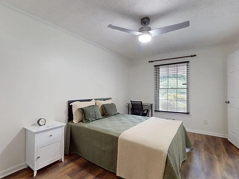 Furnished bedroom with a full bed, nightstand, and a dedicated desk area, plus a window with blinds and ceiling fan.