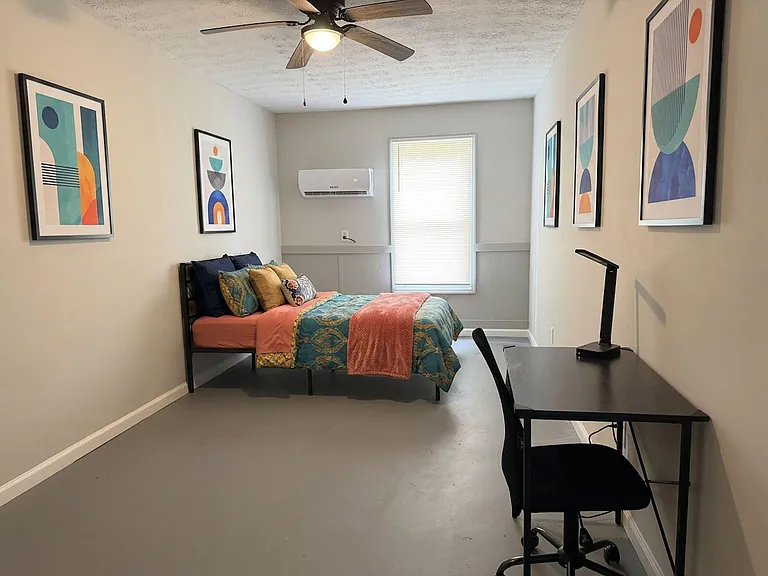 Furnished bedroom with a full bed, dedicated desk workspace, ceiling fan, and window blinds for light control.