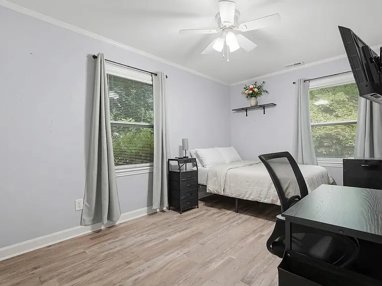 Spacious bedroom with two windows, ceiling fan with light, a work desk setup, and wall-mounted TV for easy relaxation.