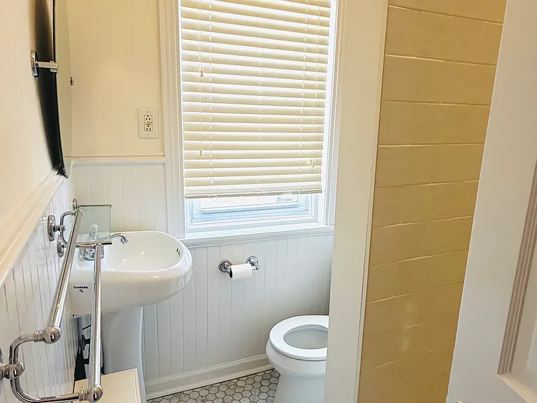 Pedestal sink with chrome fixtures, frameless oval mirror, glass shelf, and towel bars against beadboard paneling.