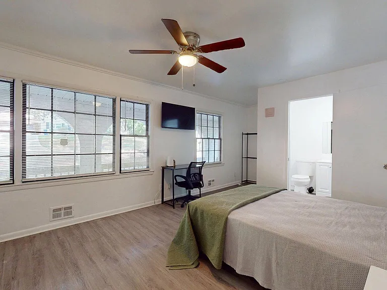 Spacious bedroom with large windows, ceiling fan, wall-mounted TV, and a desk workspace; ensuite bath access.