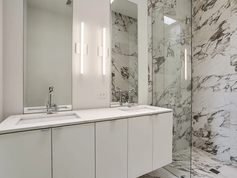 Dual vanity with integrated sinks, white cabinetry, contemporary wall-mounted lighting, large frameless mirrors, and a glass-enclosed shower featuring full-height marbled stone walls and flooring