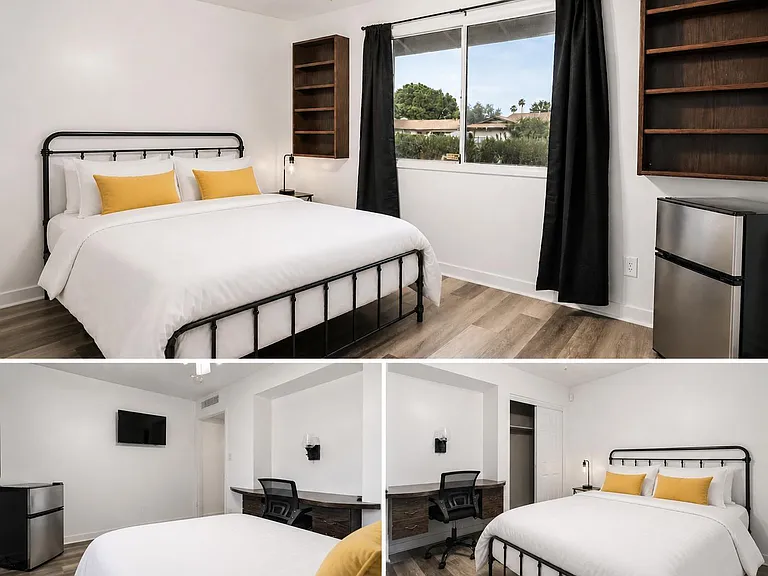Furnished bedroom with a full bed, workstation, wall shelving, mini fridge, and a large window with blackout curtains.