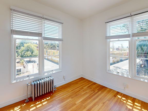 Bright room featuring large windows with blinds, hardwood floor, and a vintage radiator, overlooking a leafy street