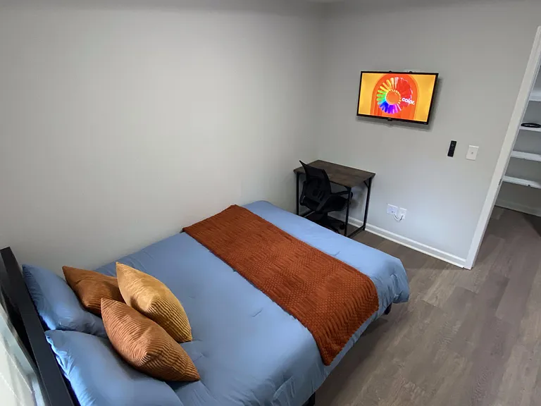 Furnished bedroom with a full bed, wall-mounted TV, and a compact desk setup, plus open shelving for storage.