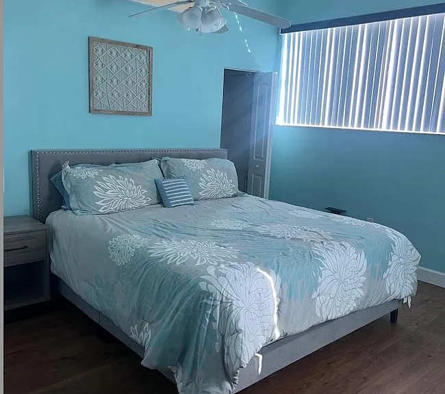 Spacious bedroom with a comfortable bed, ceiling fan, and wood-style flooring, plus a large window with vertical blinds.