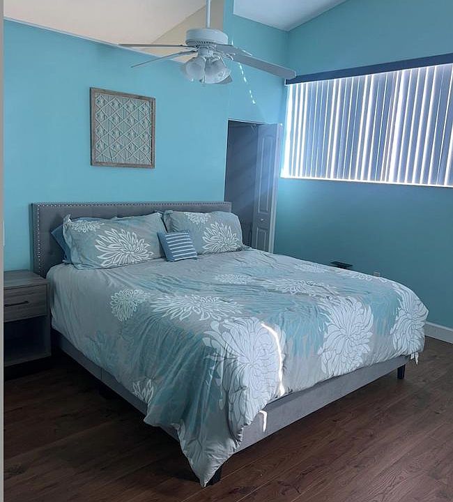 Spacious bedroom with a comfortable bed, ceiling fan, and wood-style flooring, plus a large window with vertical blinds.