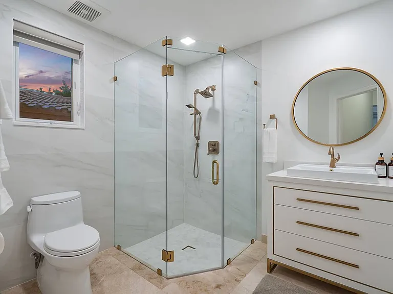 Indulge in elegance with a glass-enclosed rain shower, brass fixtures, and a chic marble vanity, set against crisp white walls.