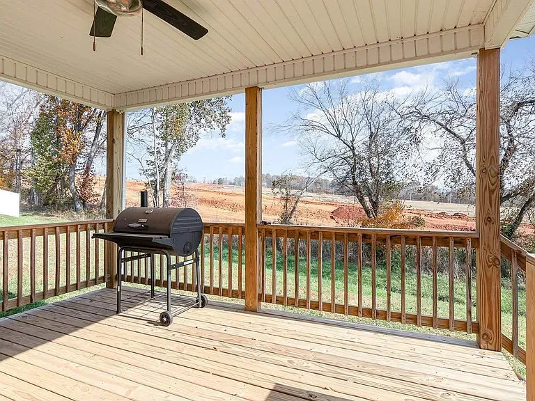 Covered back porch with ceiling fan, wood decking, and railingideal for grilling and enjoying peaceful outdoor views.