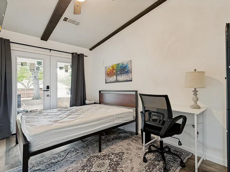 Bright furnished bedroom with private entrance through curtained double patio doors, with work desk, nightstand, & wardrobe