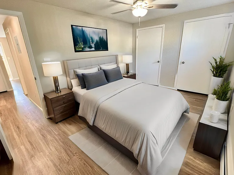 Elegant bedroom with modern decor, plush bedding, stylish ceiling fan, and warm hardwood floors, enhanced by natural lighting.