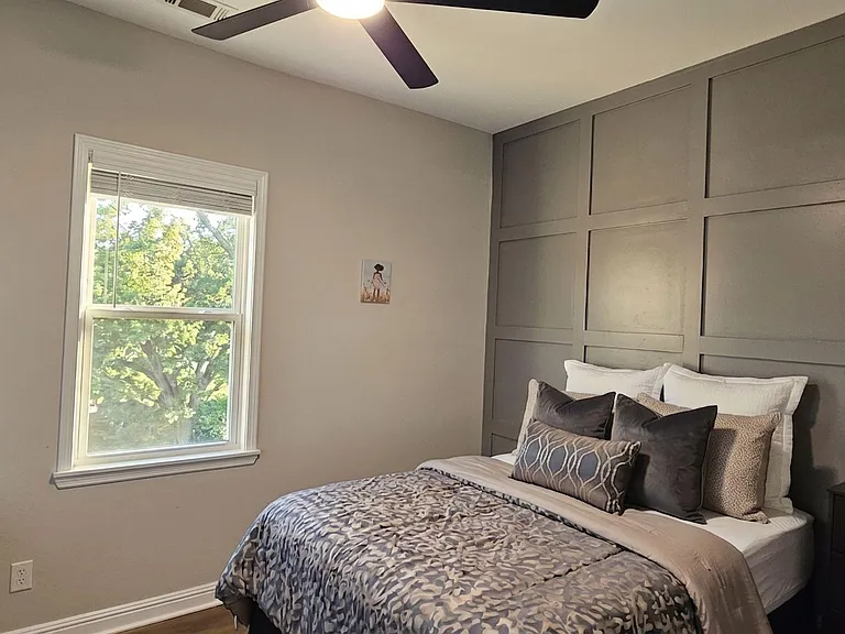Furnished bedroom with a paneled accent wall, ceiling fan, wall-mounted Smart TV, dresser, work desk with a view & 2 windows