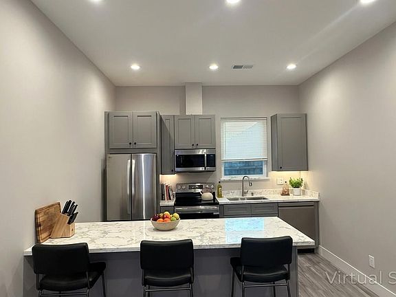 Bright and open brand new kitchen with a generous quartz breakfast bar, stainless steel appliances, gray cabinets, and recessed lighting. Virtual AI staging shown for illustrative purposes only.