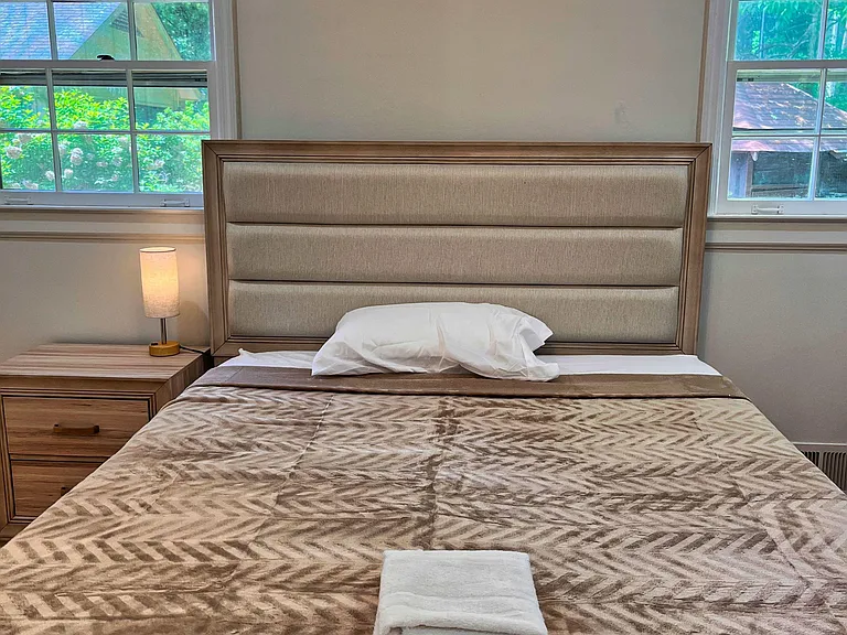 Furnished bedroom with a comfortable bed, nightstand lighting, and two windows offering leafy views and natural light.