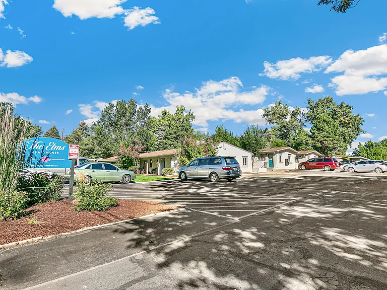 Charming Elms Apartments with spacious parking and lush greenery under a bright blue sky.