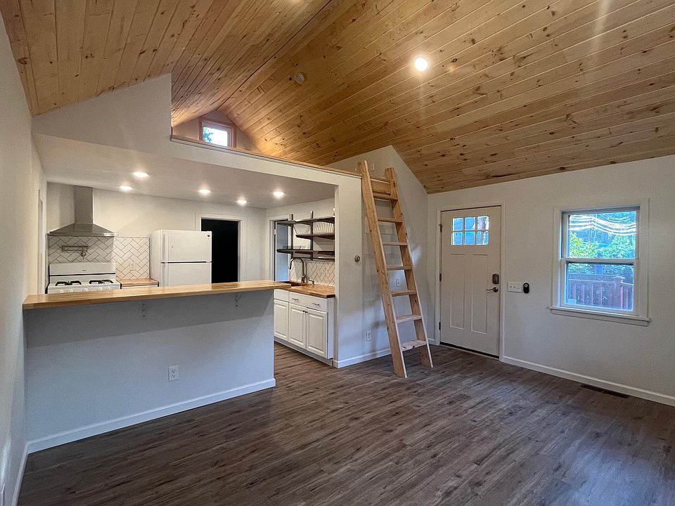 view from living room. includes front door, ladder to loft, kitchen with peninsula with bar seating. door to right is to bathroom and door straight back is to bedroom