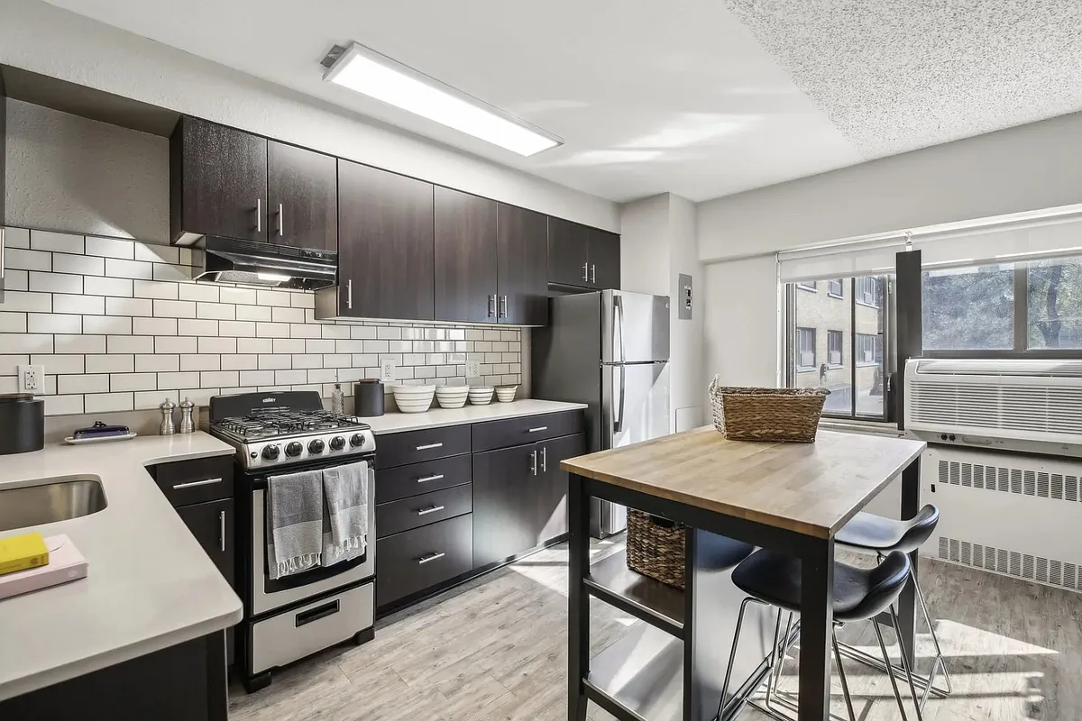 This kitchen features black cabinets and white appliances, as well as a wooden table - Archer Tower