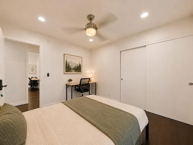 Furnished bedroom with a full bed, dedicated work desk, ceiling fan, and large sliding-door closet for storage.