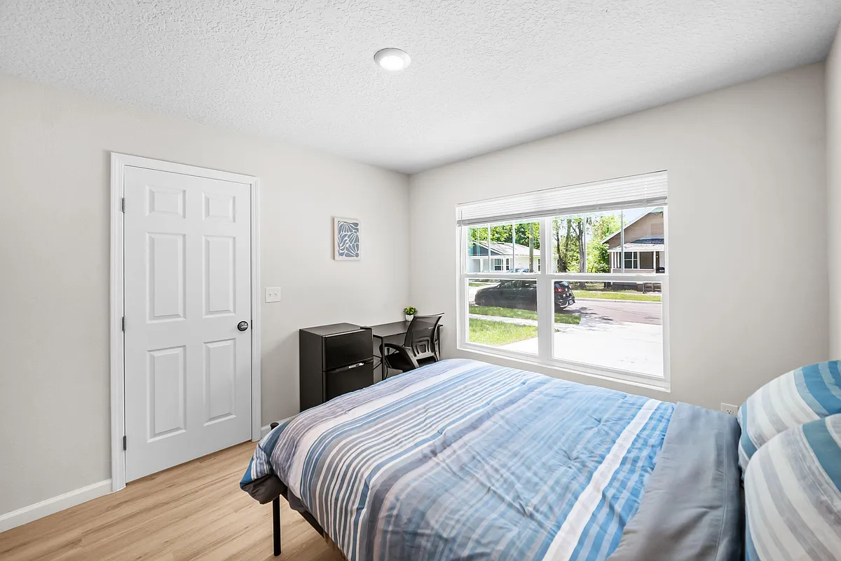 Well-sized bedroom with a large window, comfortable bed, and a dedicated desk area plus compact mini fridge for convenience. - E 18th St
