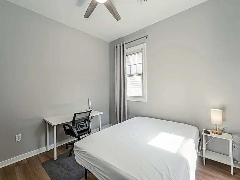 Comfortable bedroom with a furnished workspace, ceiling fan, and window with curtains for natural light and privacy.