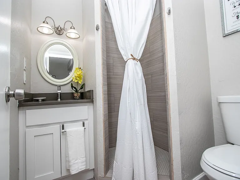 Detached casita bathroom at, Tempe rental home. Features a white vanity, an oval mirror with classic vanity lighting, a walk-in tiled shower with a white curtain, and a clean toilet area. Bright and functional bathroom for a top Tempe, AZ stay