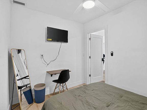 Bedroom with a ceiling fan, wall-mounted TV, and a compact wall desk setup, plus wood-look flooring and a full-length mirror.