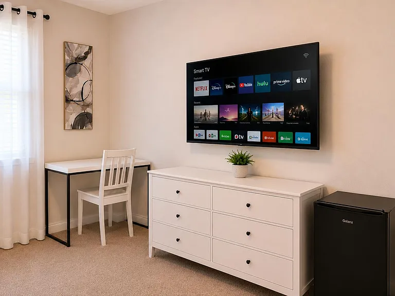 Furnished room with a dedicated desk area, dresser storage, wall-mounted smart TV, and in-room mini fridge near a window.