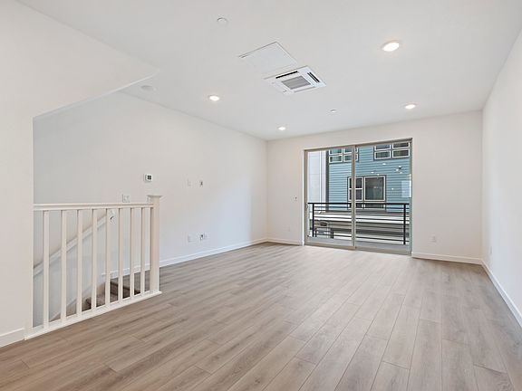A bright, open room with white walls, a wooden floor, and a balcony visible through a glass door in