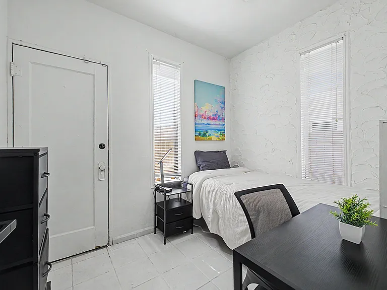 Private furnished bedroom with a full bed, desk workspace, and dresser, plus two windows with blinds for natural light.