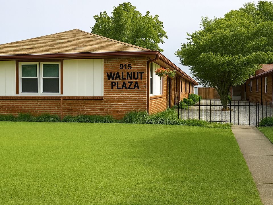 This image shows Walnut Plaza, a single-story red brick apartment complex with neatly trimmed green grass in the foreground. The building features white siding accents, a bold black "915 WALNUT PLAZA" sign on the brick wall, and a courtyard with white gra