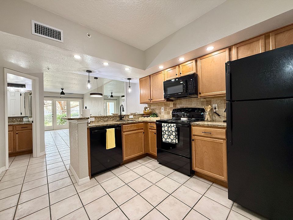 This renovated kitchen features modern black appliances, granite countertops, wood cabinetry, and pendant lighting. The open layout flows into a bright living space with tile flooring and French doors leading to the patio.