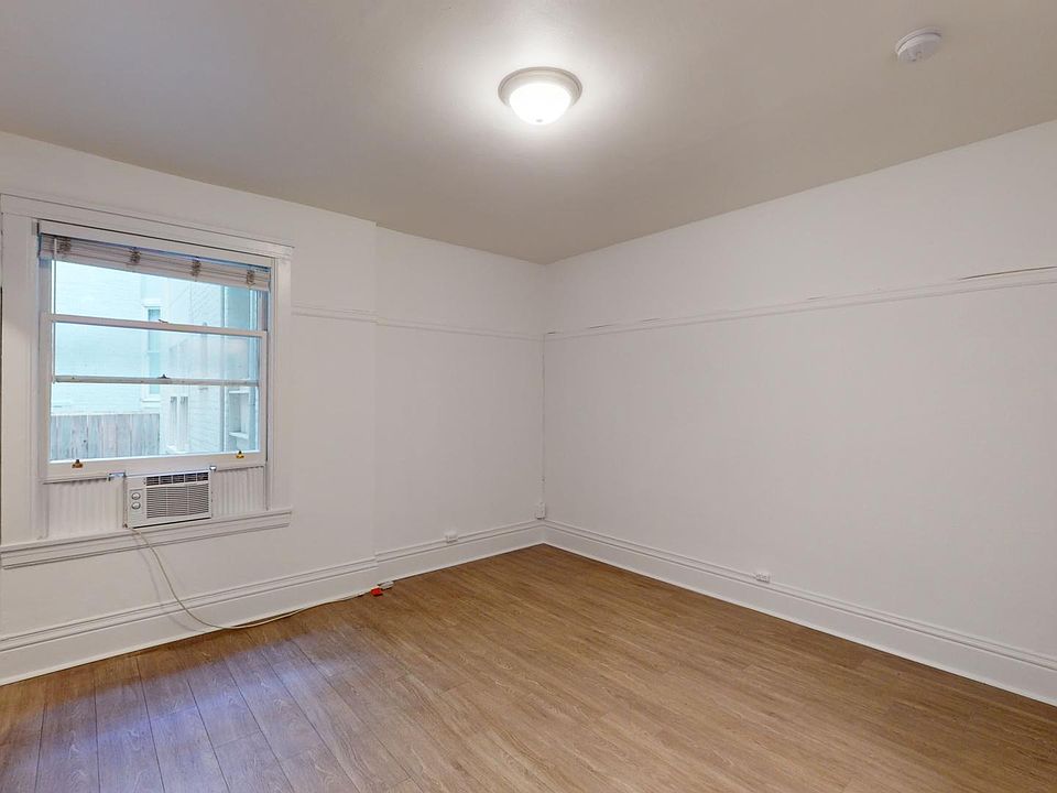 A vacant room with a wooden floor. A single window with a closed blind and an air conditioner is on the left wall. The room is well-lit with a ceiling light.