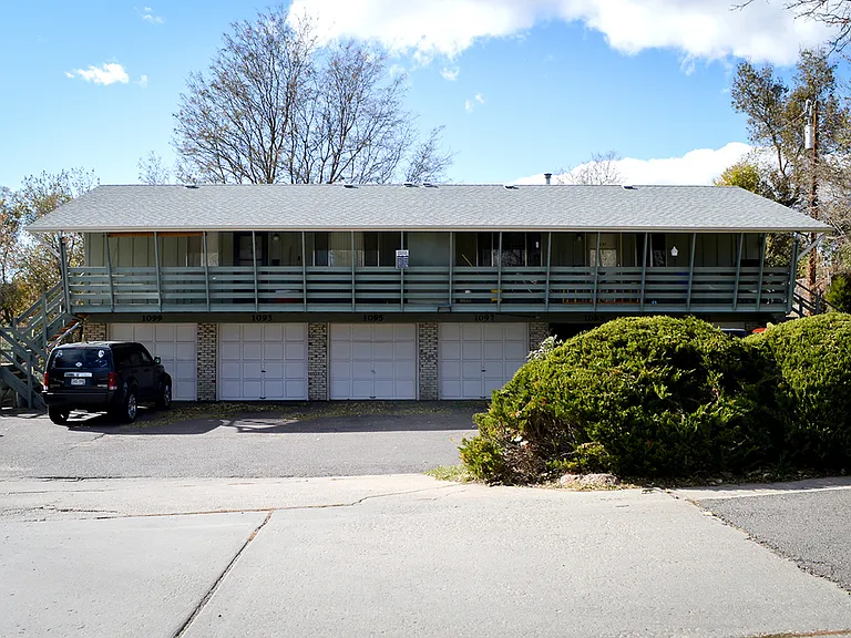 Front view of a multi-unit residential building with garages and a balcony, surrounded by greenery.