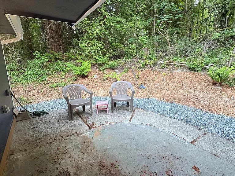 Covered patio with seating and a concrete pad, opening to a private wooded yard with low-maintenance gravel paths.