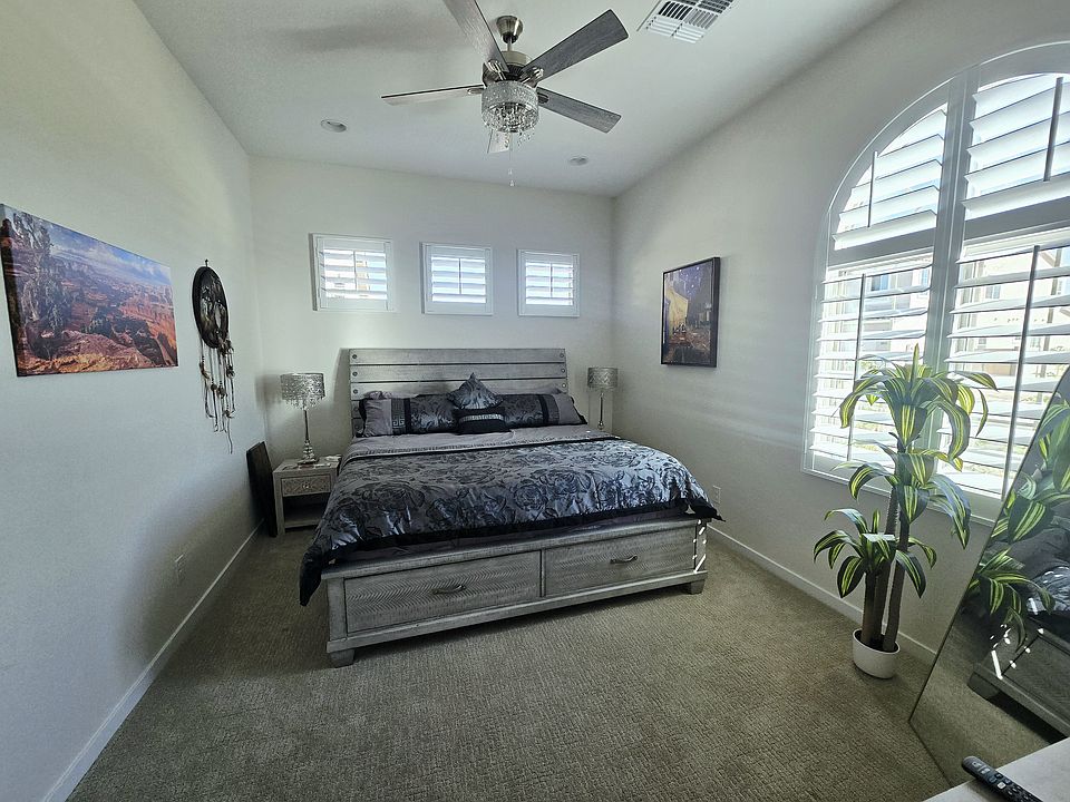 Spacious bedroom with wood bed frame and built-in drawers, matching nightstands, and lamps that complement the chandelier fan. Dresser offers extra storage. Tall mirror and faux plant sit near arched window with wood shutters. Soft light and calm decor.