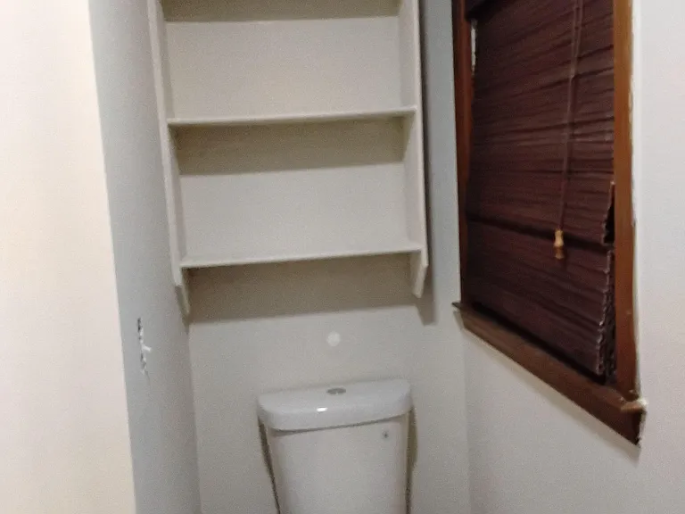 Compact bathroom area with a toilet, window with blinds, and built-in wall shelving for linens and toiletries.