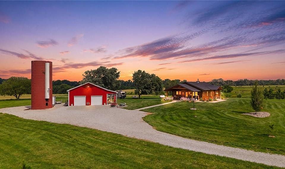 Expansive property featuring a gravel driveway, a red barn with two garage doors, a tall red silo, a single-story residence with a covered porch, and extensive manicured lawns